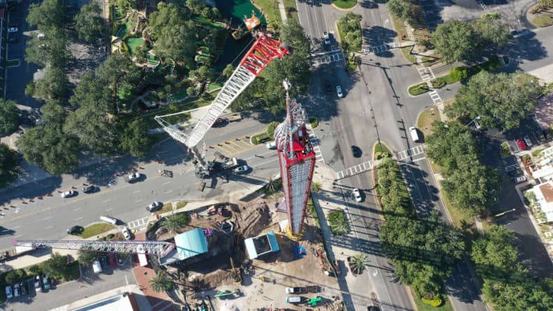 Bird’s-eye view of the future site for a record-breaking thrill ride, where cranes and crews are at work on a bustling intersection lined with trees and passing cars, setting the stage for what’s anticipated to be the world’s tallest drop tower.