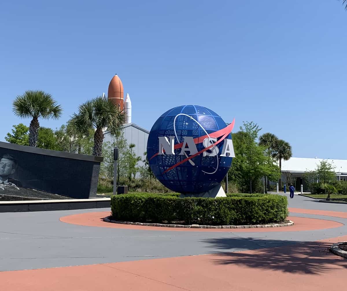 The impressive NASA globe welcomes guests at the Kennedy Space Center Visitor Complex, surrounded by palm trees and a towering rocket, all set against a vibrant blue sky—a must-see photo spot for space fans where the spirit of real astronauts inspires adventure and imagination.