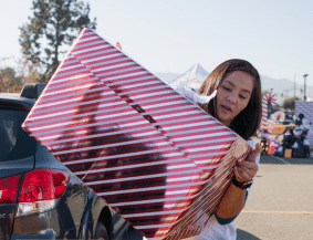 A Disney fan carries a large, red-and-white striped gift box outside near parked cars and trees during the Ultimate Toy Drive, joining in the magic of giving.