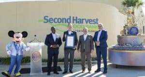 Mickey Mouse joins four Disney team members outside Second Harvest Food Bank, holding a framed certificate beside a statue of Walt Disney and Mickey.