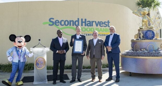 Mickey Mouse joins four Disney team members outside Second Harvest Food Bank, holding a framed certificate beside a statue of Walt Disney and Mickey.