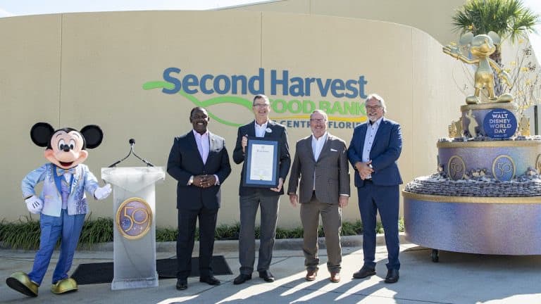 Mickey Mouse joins four Disney team members outside Second Harvest Food Bank, holding a framed certificate beside a statue of Walt Disney and Mickey.