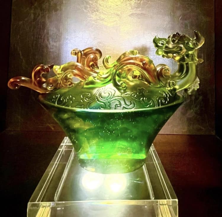 The green and amber glass dragon sculpture, inspired by Nine Dragons Restaurant at EPCOT, glows on its illuminated display stand against a dramatic dark backdrop, adding a magical touch to the World Showcase atmosphere.