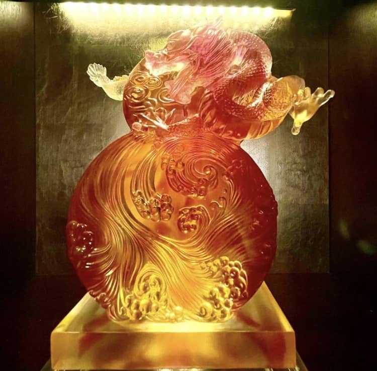 A dazzling amber dragon sculpture, echoing the enchanting decor at Nine Dragons Restaurant in EPCOT’s World Showcase, coils majestically around an intricate, swirling orb and glows with dramatic overhead lighting.
