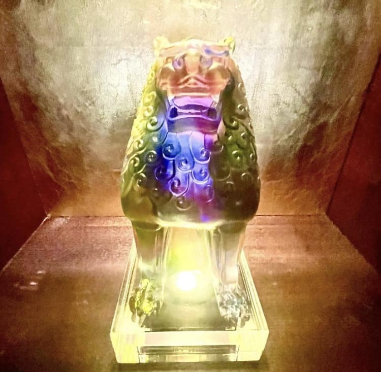 A vibrant glass lion statue on a square pedestal, illuminated from below and set against a textured brown backdrop—this dazzling piece mirrors the lively, immersive decor of Nine Dragons Restaurant at EPCOT’s China Pavilion.