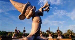 Magical stone statue of Mickey Mouse as the Sorcerer’s Apprentice from Fantasia, set against lush trees and a bright blue sky in the heart of a Disney park.