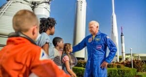 A Kennedy Space Center astronaut in a blue suit greets kids at the NASA rocket display under sunny skies, creating an exciting interactive experience for visitors.