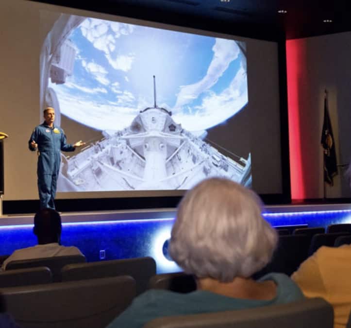 A Kennedy Space Center presenter in a blue suit delivers an engaging talk in the auditorium, with an impressive image of Earth from space lighting up the big screen behind them.