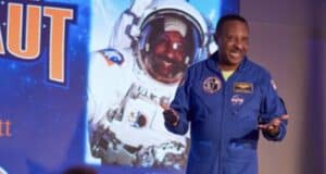 A NASA astronaut takes the stage at Kennedy Space Center Visitor Complex, set against a dramatic backdrop of outer space imagery and an illuminated "ASTRONAUT" sign.