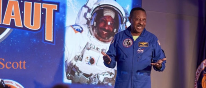 A NASA astronaut takes the stage at Kennedy Space Center Visitor Complex, set against a dramatic backdrop of outer space imagery and an illuminated "ASTRONAUT" sign.