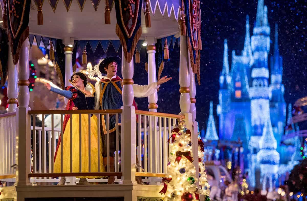 Snow White and her prince greet fans from a dazzling parade float at Disneyland, waving as Sleeping Beauty Castle glows in the night behind them and the park sparkles with enchanting magic.