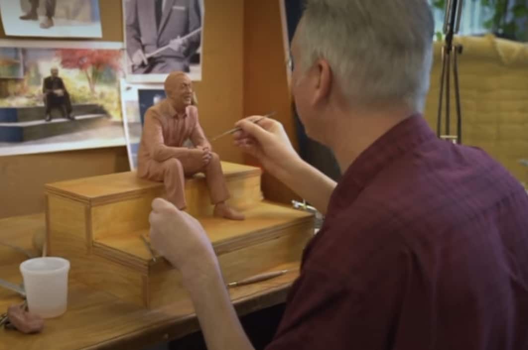 In a creative studio buzzing with Disney magic, an Imagineer shapes a clay sculpture of a seated character, drawing inspiration from reference photos and historic sketches of Walt Disney himself pinned on the wall—a nod to the innovative artistry behind beloved attractions.
