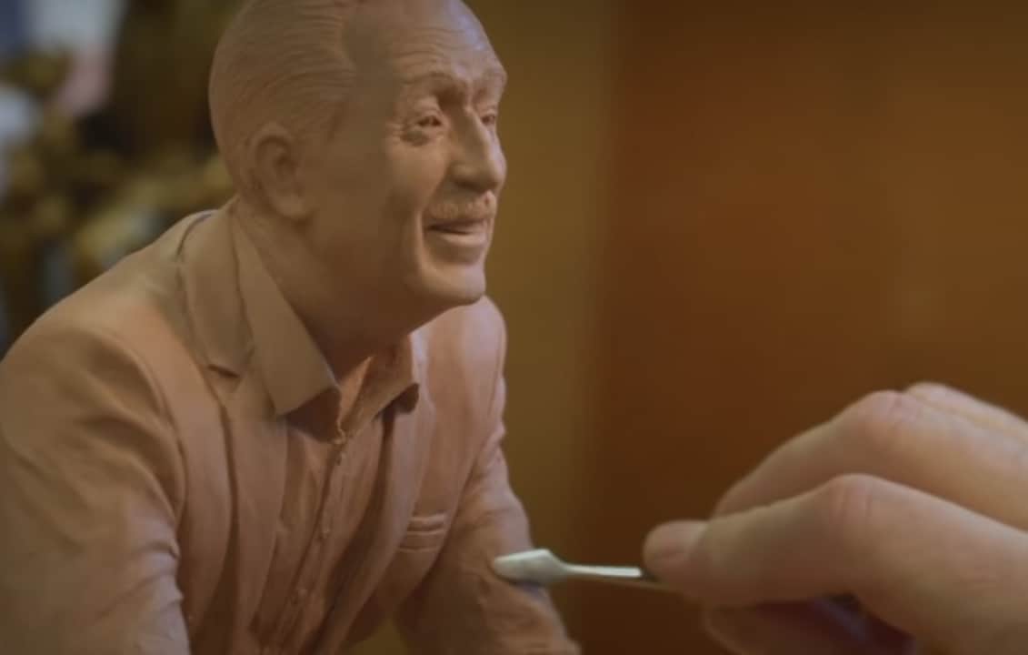 An artist skillfully shapes a clay bust of a cheerful man, using fine tools to capture intricate shirt details inspired by Walt Disney’s classic look—a creative process that brings Disney magic to life before your eyes.