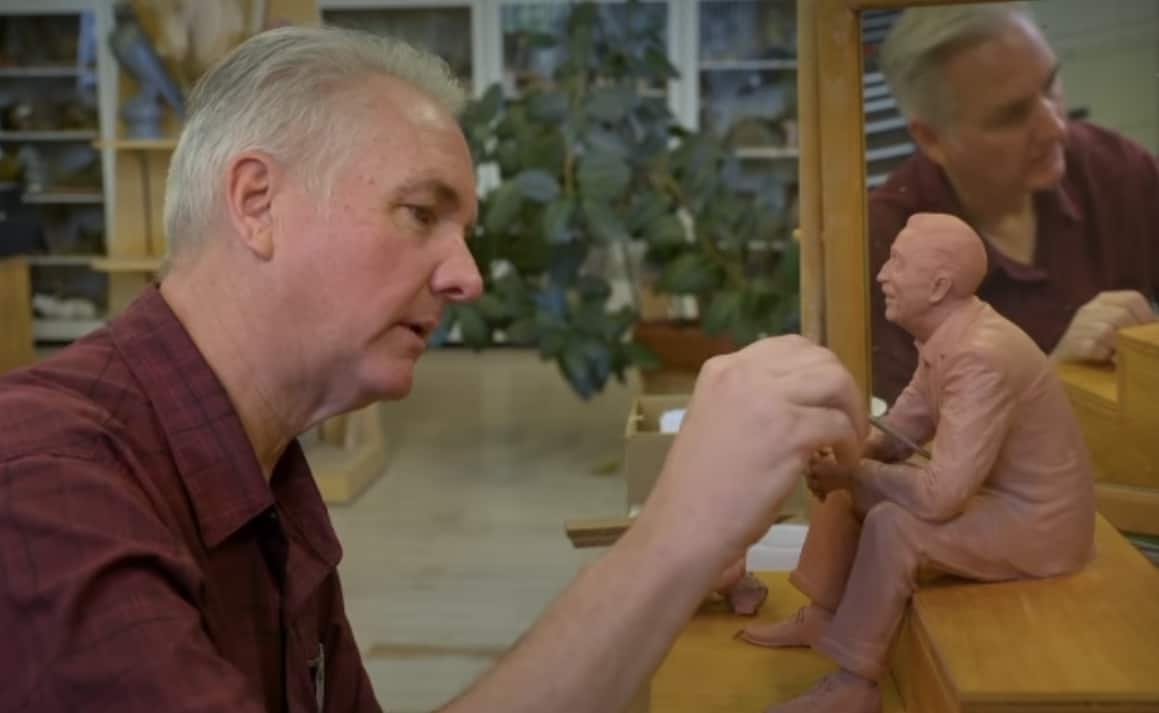 An artist in his studio meticulously sculpts a seated figure, with a mirror reflecting his progress—a creative scene reminiscent of the workshops where Walt Disney and Imagineer Zach Riddley shaped their legendary theme park masterpieces.