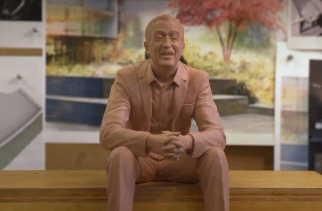 A detailed clay sculpture of Walt Disney in a suit sits on a bench, surrounded by imaginative artwork and lush trees, capturing the creative energy that inspired Disney Parks and honoring both Walt Disney and Imagineer Zach Riddley.