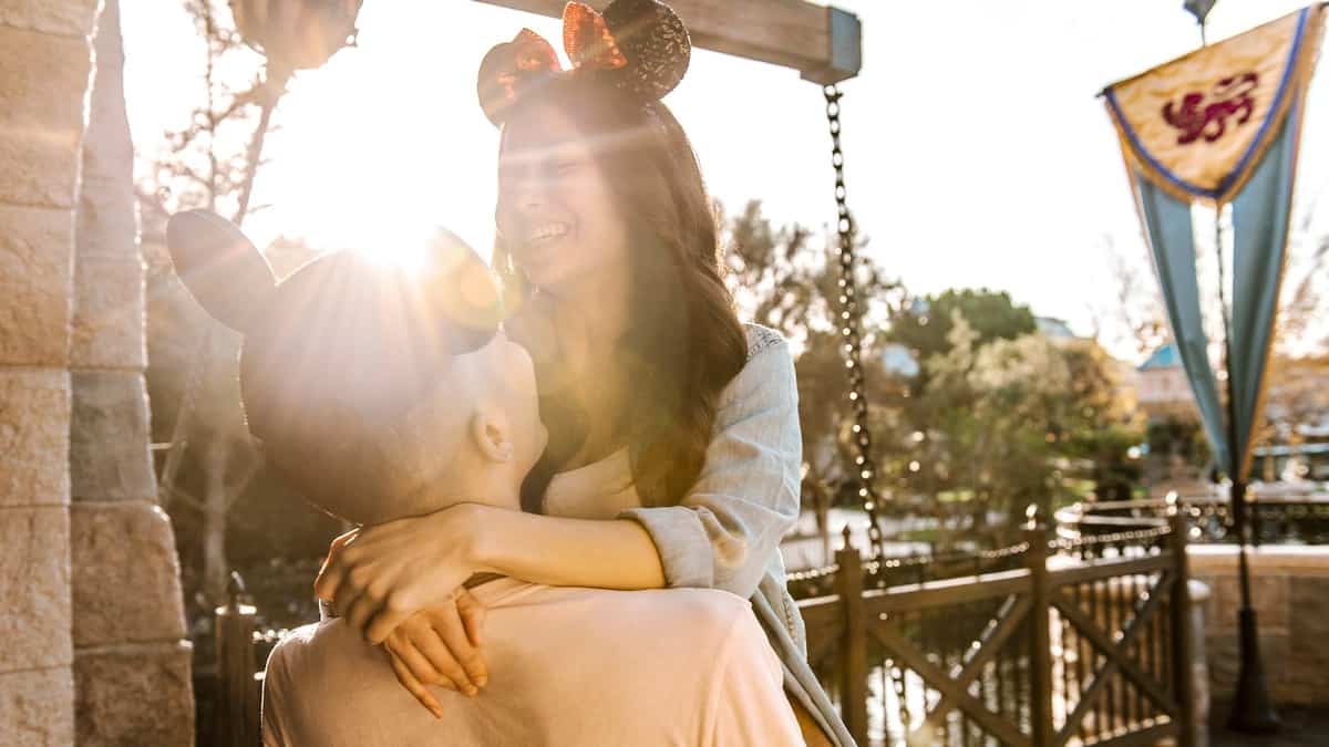 Disneyland guests wearing mouse ears embrace and smile beneath the sun, surrounded by festive banners and lush trees, evoking the romance and excitement of Sweethearts’ Nite during Disneyland After Dark.