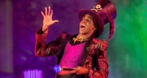 A charismatic Disneyland performer in a vibrant, eye-catching costume and tall hat grins on stage, holding a glowing orb during Villains Nite at Disneyland After Dark—immersing guests in an enchanting evening packed with unique villain encounters and exclusive event offerings.