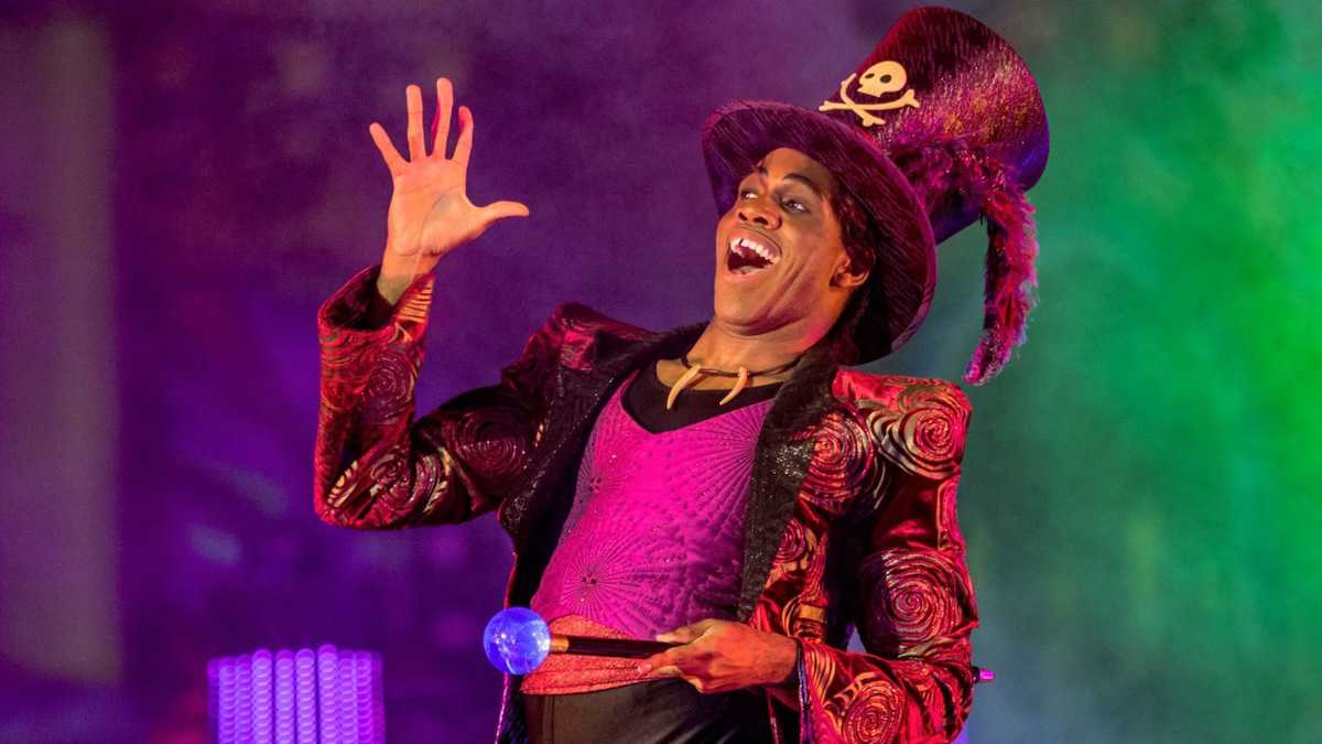 A charismatic Disneyland performer in a vibrant, eye-catching costume and tall hat grins on stage, holding a glowing orb during Villains Nite at Disneyland After Dark—immersing guests in an enchanting evening packed with unique villain encounters and exclusive event offerings.