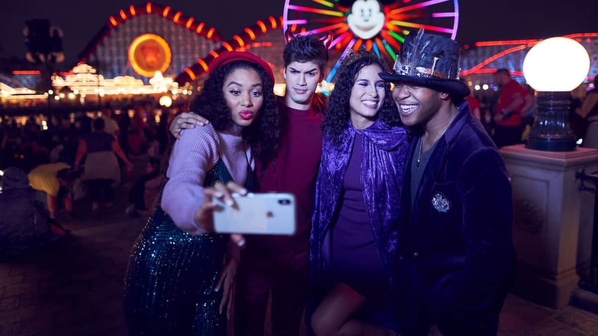 Four friends in themed outfits laugh and take a group selfie amid the vibrant lights of Disneyland After Dark: Villains Nite, immersing themselves in the event’s thrilling atmosphere at Disneyland after hours.