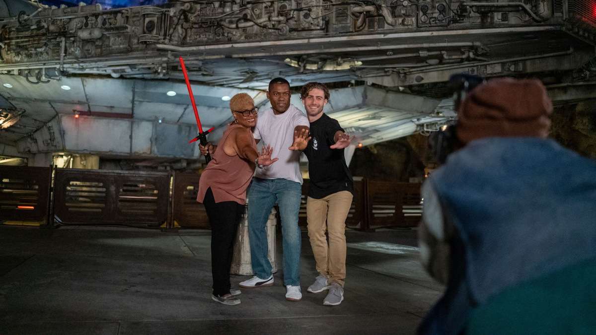Fans strike playful poses in front of a towering Star Wars spaceship at Disneyland After Dark: Star Wars Nite, while someone captures the fun with a nighttime photo amidst the park’s vibrant atmosphere.