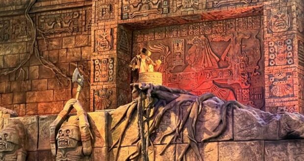 A gleaming golden idol stands atop a pedestal in front of an ancient-looking, vine-draped stone wall adorned with intricate carvings—an immersive scene that feels straight out of an Indiana Jones Adventure attraction at a Disney park, capturing the thrilling spirit found in 2021 Disney movie settings.