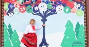 A cheerful youngster in a vibrant red skirt and crisp white shirt strikes a pose beside a lively holiday mural featuring joyful cartoon characters and sparkling decorated trees, capturing the festive spirit of the park’s seasonal celebration.