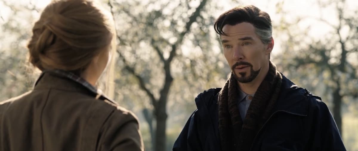 Two guests chat in a softly lit area of an immersive Disney park; one smiles at the camera while the other, partially hidden among enchanted trees, brings to mind a scene plucked from a Marvel Studios teaser like Doctor Strange in the Multiverse of Madness.