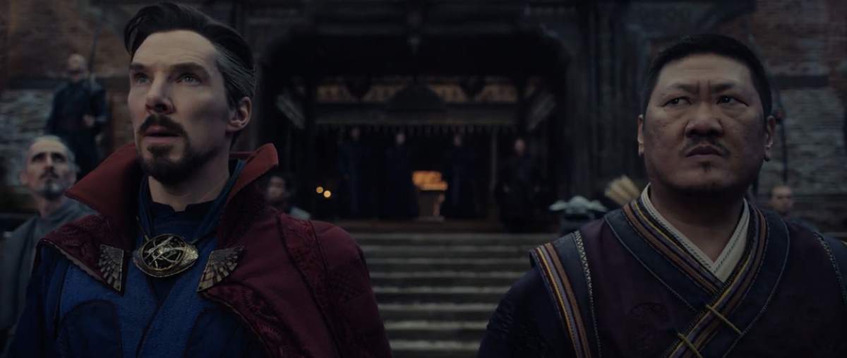 Doctor Strange and Wong, dressed in their signature capes, stand side by side with serious expressions on the stone steps of Avengers Campus, as park guests pass by in the background—capturing that teaser-trailer vibe from Doctor Strange in the Multiverse of Madness.