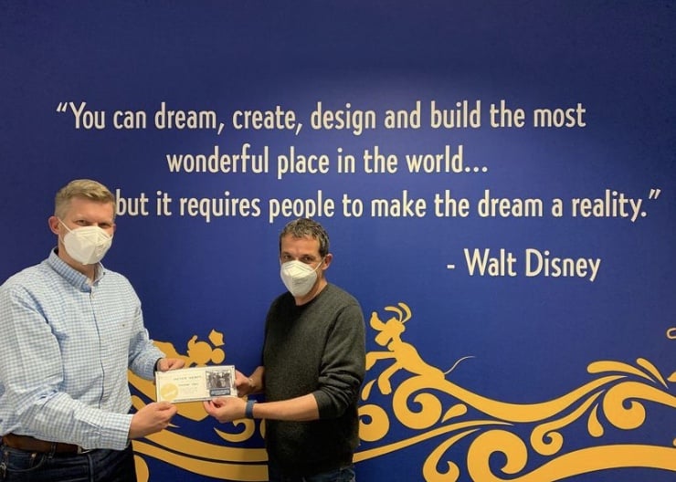 Disney Wish shipyard team members, wearing masks, proudly hold up an official document in front of a famous Walt Disney quote on a blue wall, marking an exciting milestone for Disney Cruise Line.