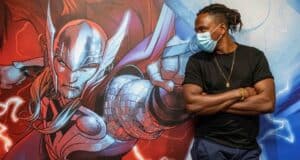 A masked Marvel fan poses confidently with arms crossed beside a vibrant comic-style Thor mural, capturing the excitement of a superhero attraction.