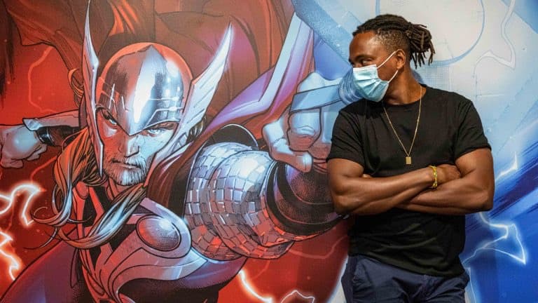 A masked Marvel fan poses confidently with arms crossed beside a vibrant comic-style Thor mural, capturing the excitement of a superhero attraction.