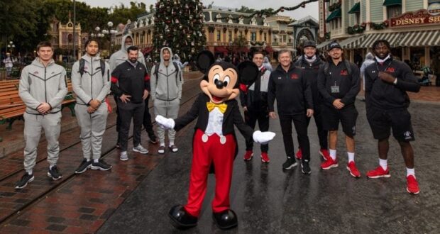 Mickey Mouse joins guests dressed in sporty outfits for a festive photo op by the sparkling Christmas tree at Disneyland, spreading holiday cheer throughout the park.