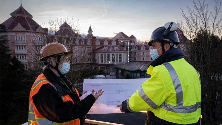 Cast Members in high-visibility vests and hard hats examine blueprints outside the Disneyland Hotel Paris at sunset, gearing up for the much-anticipated Royal Makeover of this iconic Disney landmark.