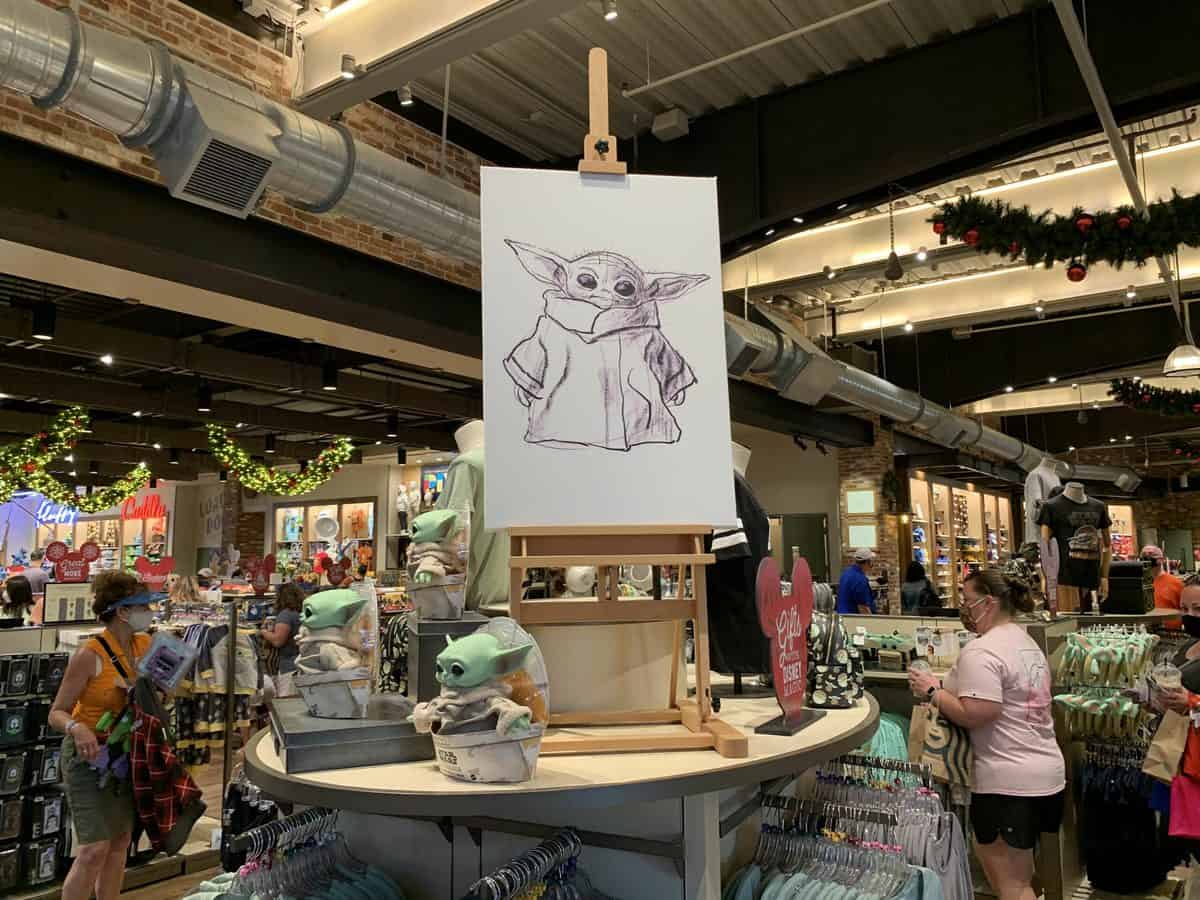 A lively Star Wars gift shop buzzes with fans admiring a creative Baby Yoda sketch displayed on an easel, surrounded by shelves filled with Grogu merchandise as guests rush to check out and make the most of their theme park visit.