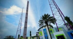 Experience the adrenaline rush as you gaze up at Funland’s record-breaking World's Tallest Drop Tower and the heart-pounding Slingshot, soaring above vibrant park buildings lined with swaying palm trees, all set against a partly cloudy sky.