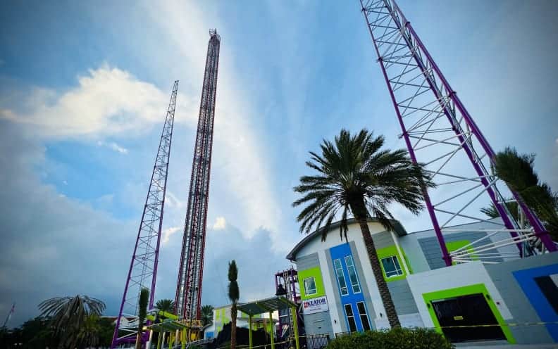 Experience the adrenaline rush as you gaze up at Funland’s record-breaking World's Tallest Drop Tower and the heart-pounding Slingshot, soaring above vibrant park buildings lined with swaying palm trees, all set against a partly cloudy sky.