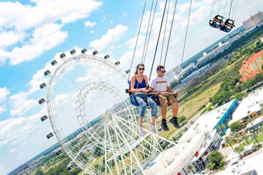 Thrill-seekers enjoy the Skyscreamer swing ride at Six Flags, gliding high above the park with the towering Ferris wheel and city skyline as their backdrop, soaring past Lex Luthor: Drop of Doom—the tallest drop tower in operation today.
