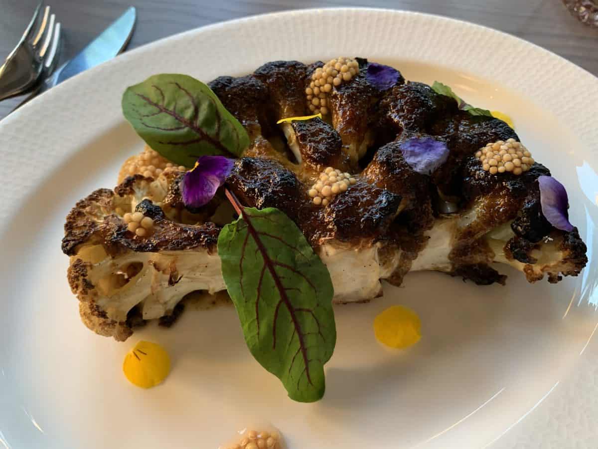 A colorful roasted cauliflower steak topped with fresh greens, mustard seeds, and purple flower petals sits on a white plate, channeling the playful flair and imaginative flavors you’d find at Four Flamingos in Orlando—a dish that could easily be served at an EPCOT food festival for fans of Richard Blais’ creative cuisine.