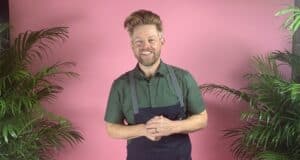A cheerful staff member in a vibrant green shirt and apron stands between lush plants against a bright pink backdrop, channeling the playful, tropical ambiance guests love at Four Flamingos Florida Kitchen by Richard Blais.