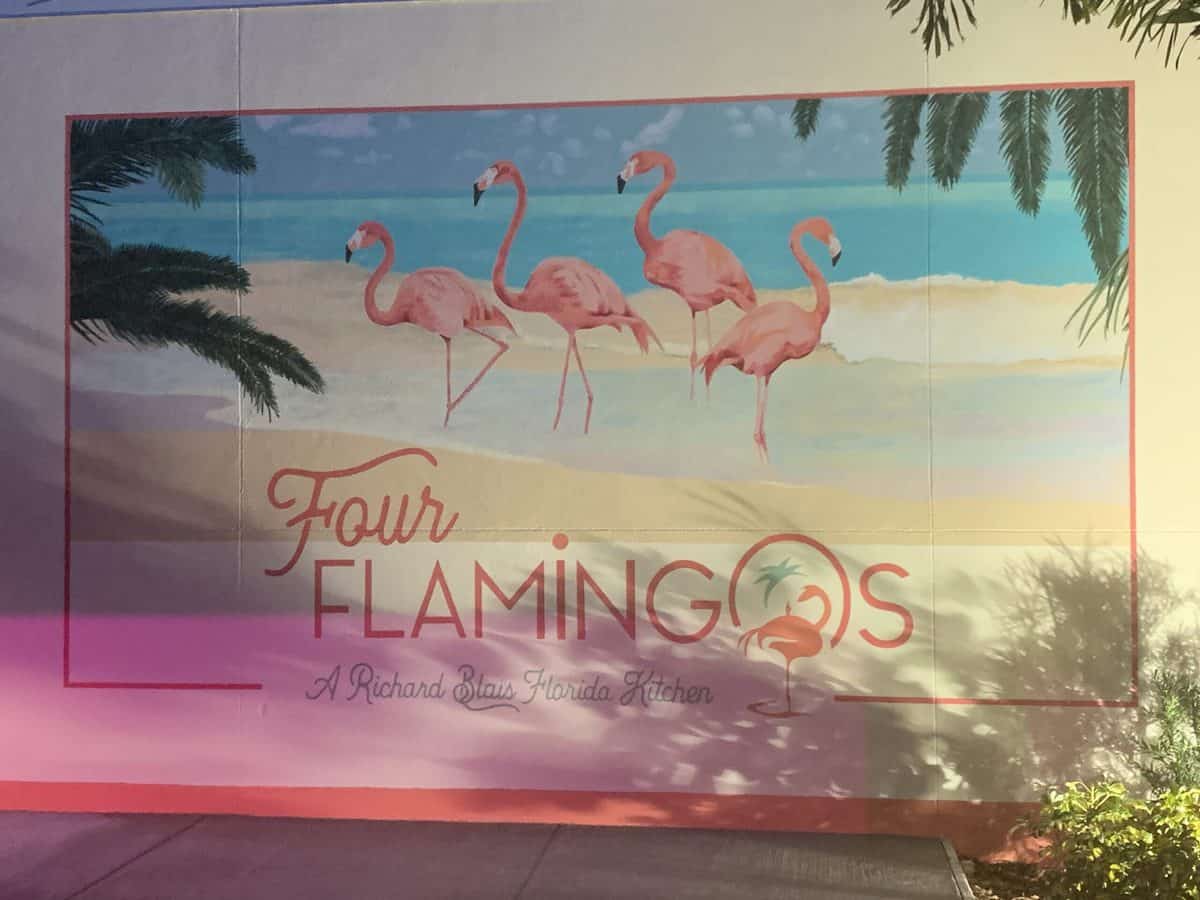 Colorful mural featuring four whimsical flamingos on a sandy beach with lush palm trees, showcasing the words "Four Flamingos: A Richard Blais Florida Kitchen." The lively scene sets a fun, tropical atmosphere reminiscent of a vibrant Florida resort or amusement park dining experience.