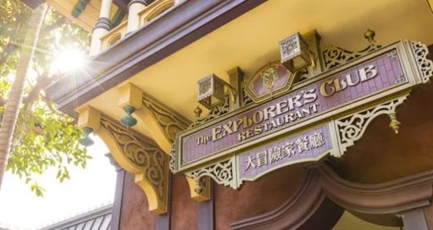 The Explorer’s Club Restaurant sign displayed on an adventurous, themed building at Hong Kong Disneyland, with sunlight streaming through lush foliage in the corner for a magical park atmosphere.