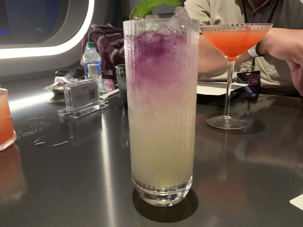Colorful, space-themed cocktail in a tall glass with vibrant purple and yellow layers and a mint garnish, served on a sleek dark table alongside other creative drinks at Space 220 Lounge in EPCOT—reservations highly recommended for this stellar Disney dining experience.
