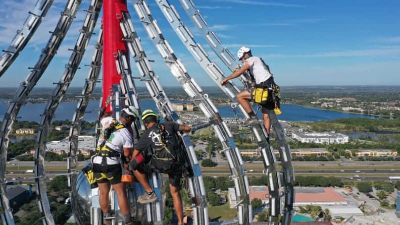 Construction crew in bright safety gear works high atop the future thrill ride, meticulously inspecting the towering steel structure that will soon set records as the world's tallest drop tower, with city buildings and speeding highway traffic far below.