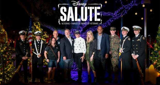 Disney veterans and military personnel gather beneath the glowing "Disney Salute" sign, surrounded by dazzling holiday lights, to celebrate during Night of a Million Lights at Give Kids The World Village.