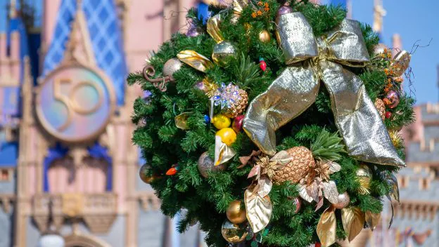 Festive holiday wreath adorned with gold ribbon and sparkling ornaments graces the foreground of Cinderella Castle at Walt Disney World, which features a prominent "50" anniversary emblem, celebrating the magical spirit of the park’s milestone season.