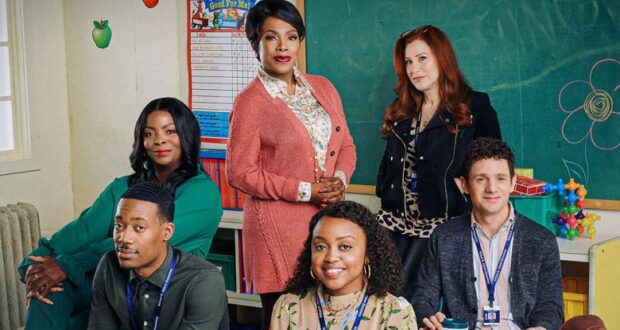 Six cast members gather in a classroom decorated with colorful children's artwork, smiling and posing together in front of a chalkboard, creating an atmosphere reminiscent of interactive theme park workshops.