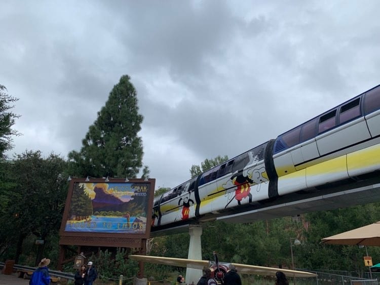 The Disneyland Monorail, featuring vibrant Mickey Mouse designs, glides past a classic park sign beneath a dramatic cloudy sky, just before the attraction temporarily closes for an exciting refurbishment.