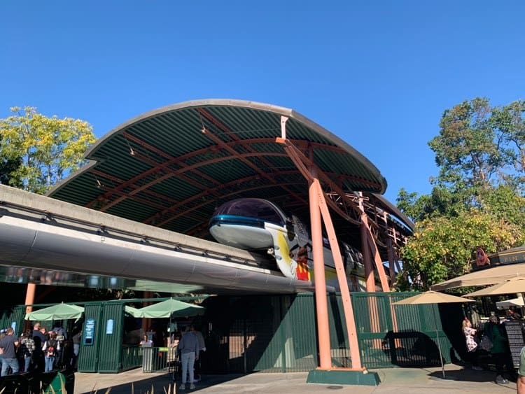 The Disneyland Monorail glides into the Tomorrowland station as fans gather under green umbrellas, enjoying the iconic ride beneath a brilliant blue sky—just days before this beloved attraction temporarily closes for refurbishment.