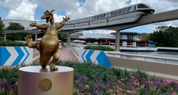 The gold dragon statue welcomes guests at EPCOT, surrounded by vibrant flowers, as a Disney monorail glides along the tracks in the background.