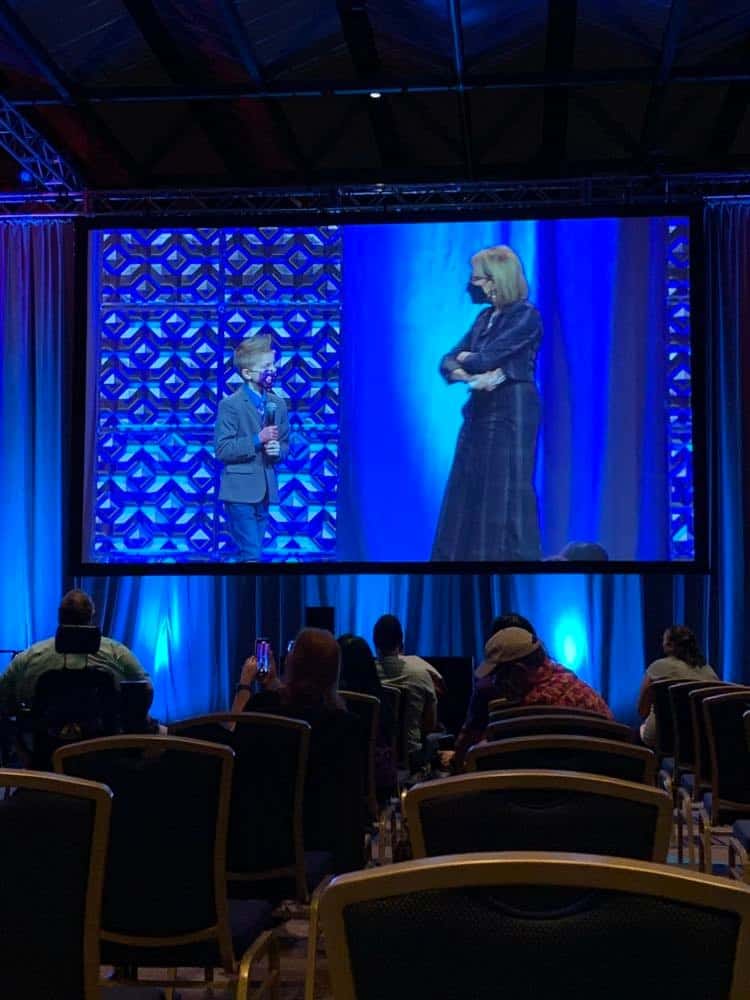 Kellen Jackley and another speaker are seen on stage, their conversation broadcast on a massive screen to a crowd of theme park and entertainment fans in a dimly lit event hall. They discuss uplifting projects like Give Kids the World, sharing real-life stories from within the industry about hope and making a difference as true World Changers.
